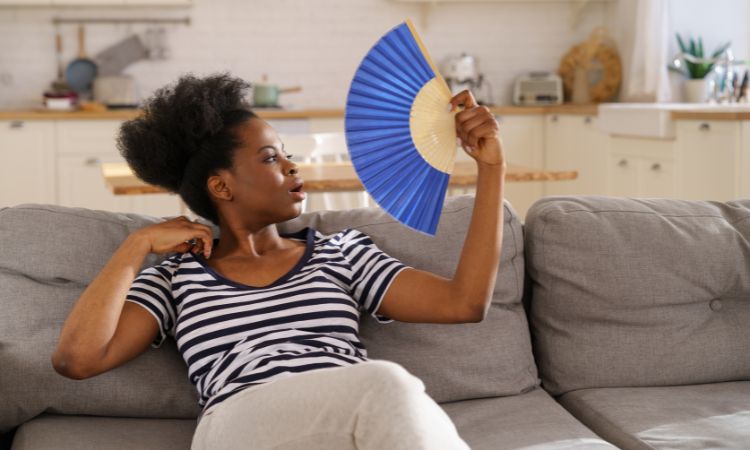 Imagen de una mujer abanicándose sentada en el sofá de su casa porque tiene mucho calor.
