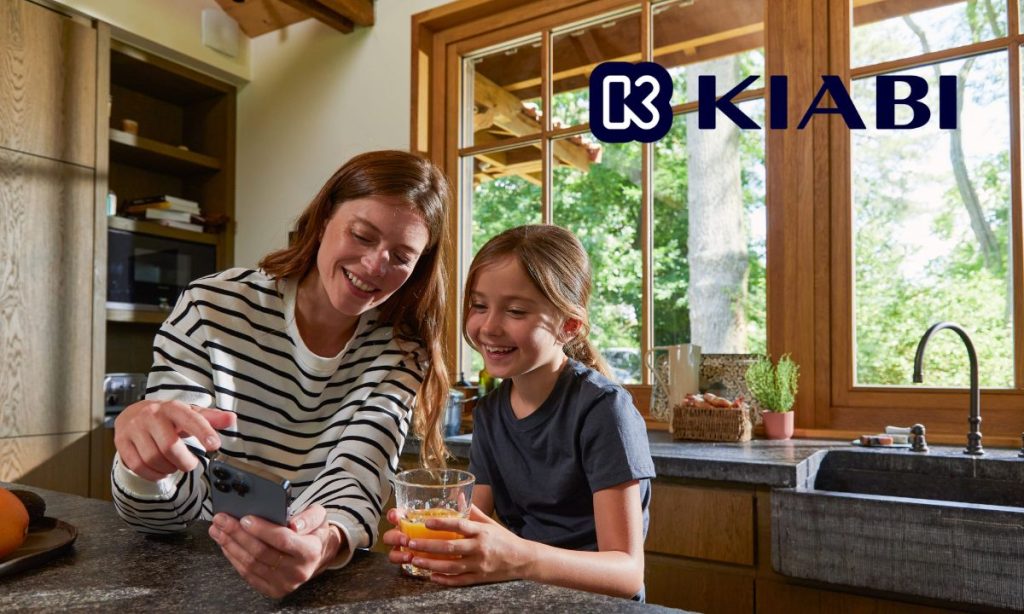 Fotografía de una madre y una hija en su cocina. Ambas están viendo productos del marketplace de Kiabi en el teléfono de la madre, sonrientes. En la esquina superior derecha de la imagen aparece el logo de Kiabi.