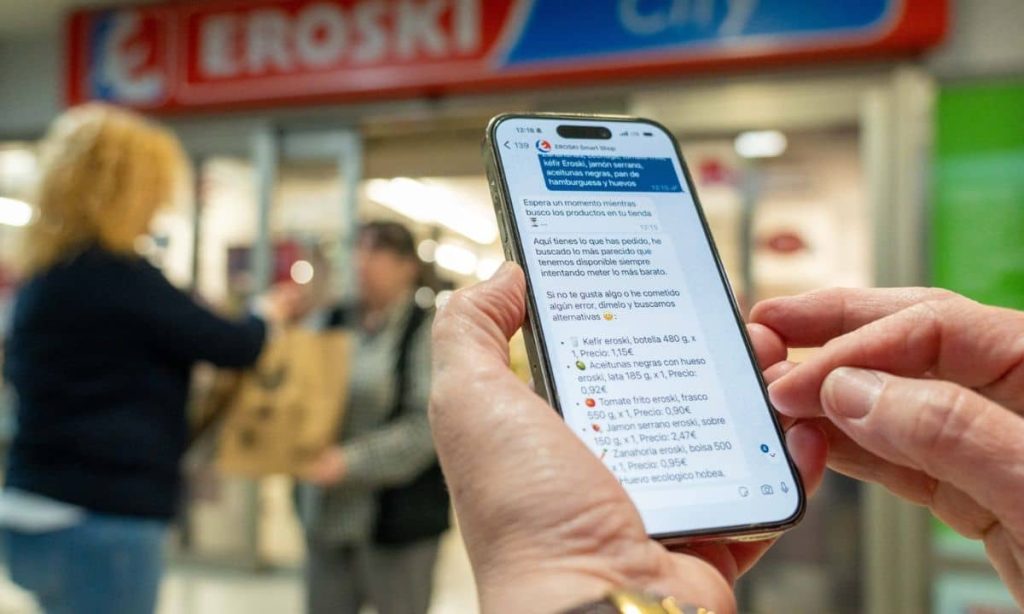 una clienta utiliza la compra por whatsapp a través de su móvil a las puertas de un supermercado eroski en el que se ve a otras dos mujeres