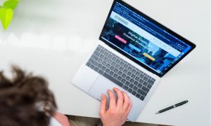 Vista cenital de una persona usando un portátil sobre un escritorio blanco, con la pantalla abierta en una web y un bolígrafo al lado.