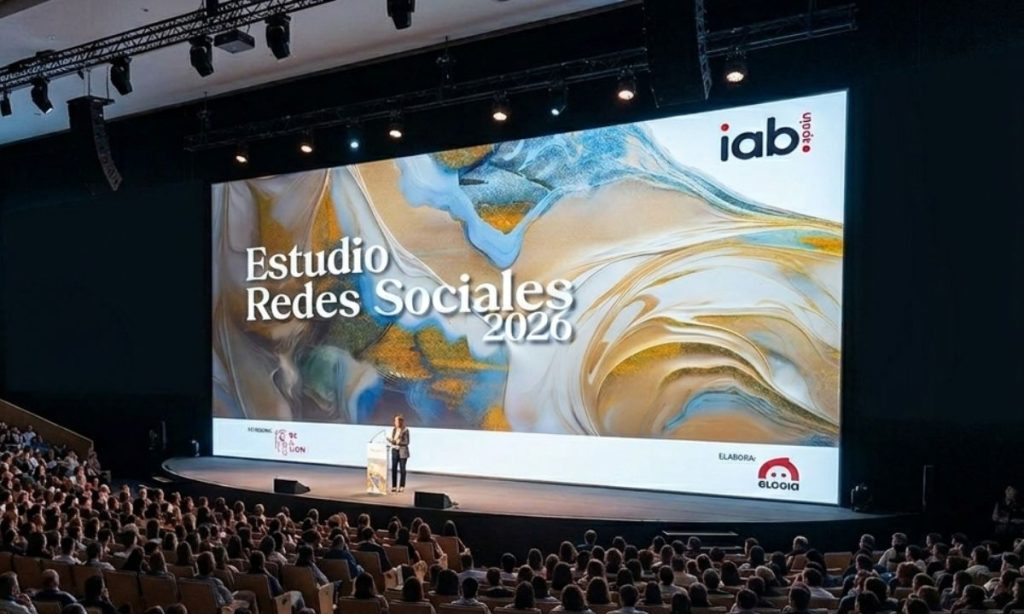 Imagen del interior de un auditorio repleto de gente. En el amplio escenario hay una persona presentando y, tras ella, se ve una pantalla gigante en la que se proyecta una creatividad de portada del Estudio Anual de Redes Sociales 2026 de IAB Spain y Elogia.