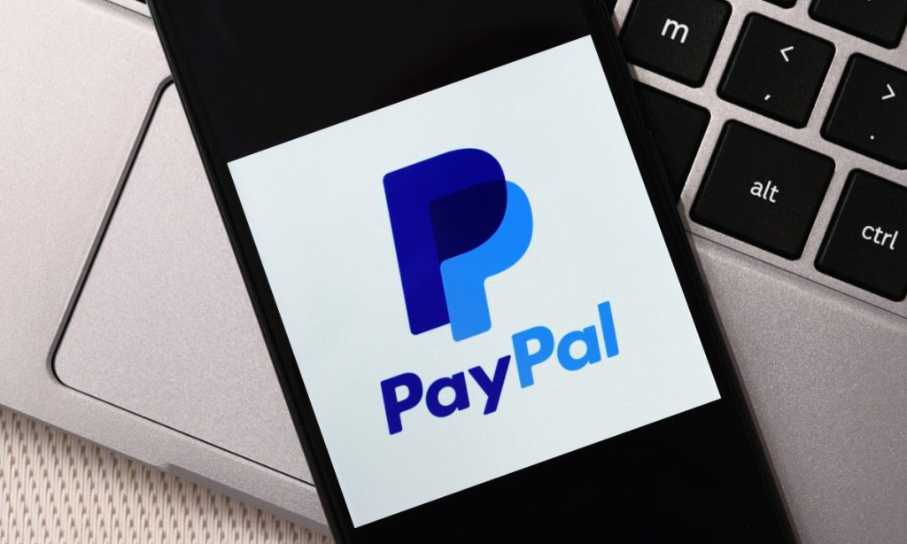 Fotografía de un móvil sobre el teclado de un ordenador portátil. En la pantalla del móvil se ve el logotipo de PayPal.