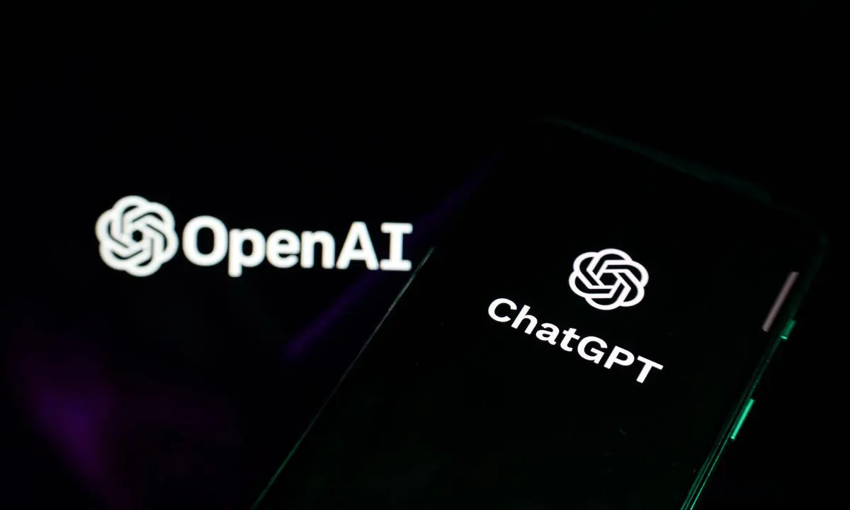 Fotografía oscura de una pantalla con el logo de OpenAI, sobre la que hay un móvil negro con el logo de ChatGPT en pantalla.